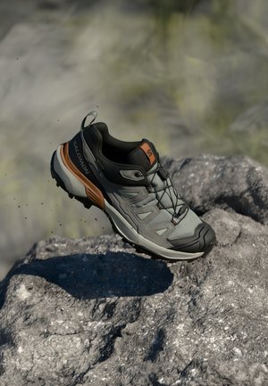 Sapatilha de trail running cinza e preta com detalhes laranja, parte superior em malha texturizada, sola de borracha e atacadores, posicionada acima de uma superfície de pedra.