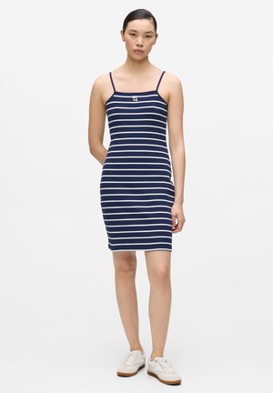 Młoda kobieta stojąca, ubrana w granatowo-białą, bezrękawnikową sukienkę w paski oraz białe trampki, na jednolitym jasnym tle.