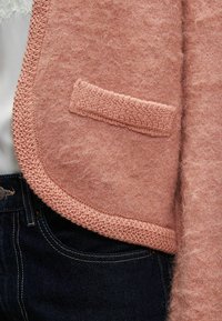 Pink fuzzy cardigan with a textured knit edge and a small pocket, worn over dark denim jeans. Soft, fluffy material, rounded hem.
