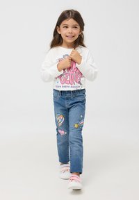 Biała koszula z długim rękawem z różowym graficznym napisem, niebieskie dżinsy z naszywkami w kształcie jednorożca i gwiazd oraz pastelowe trampki w paski.