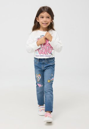 Biała koszula z długim rękawem z różowym graficznym napisem, niebieskie dżinsy z naszywkami w kształcie jednorożca i gwiazd oraz pastelowe trampki w paski.