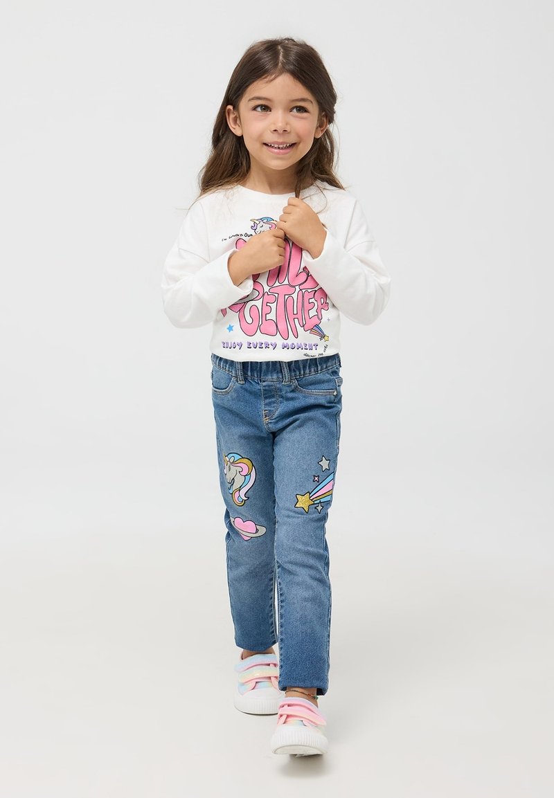 Biała koszula z długim rękawem z różowym graficznym napisem, niebieskie dżinsy z naszywkami w kształcie jednorożca i gwiazd oraz pastelowe trampki w paski.