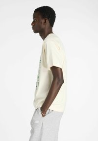 T-shirt crème avec un imprimé graphique vert, à manches courtes et à coupe décontractée, associé à un pantalon de survêtement gris. Texture en coton lisse.