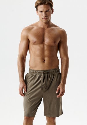 Mężczyzna bez koszulki z krótkimi blond włosami, ubran w oliwkowozielone szorty ze sznurkiem, stojący na tle jednolitego białego tła.