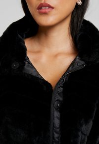 Sort faux fur jakke med lynlås foran, trykknapper og en blød, tekstureret overflade. Har høj krave og glat foring.