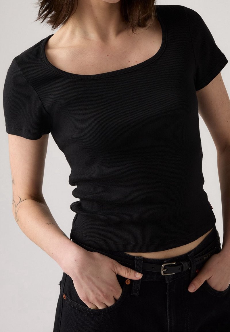 Personne portant un haut noir cintré à manches courtes avec un col rond et un pantalon noir, les mains dans les poches, sur un fond uni.