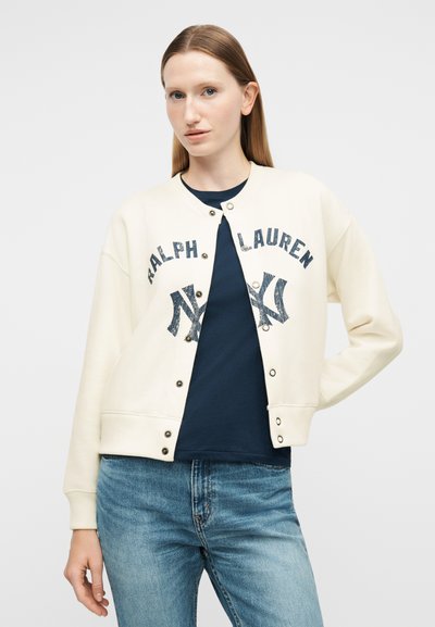 Γυναίκα που φοράει λευκό σακάκι Ralph Lauren με κουμπιά πάνω από μπλε πουκάμισο και μπλε τζιν, στέκεται με το ένα χέρι στη μέση της.