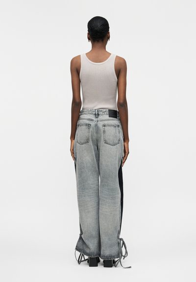 Γυναίκα με τα μαλλιά σε κότσο, που φοράει ένα λευκό αμάνικο τοπ και φαρδιά γκρίζα τζιν με μαύρες πλαϊνές ρίγες και κορδόνια στο τελείωμα, στέκεται γυρισμένη προς τα πίσω.