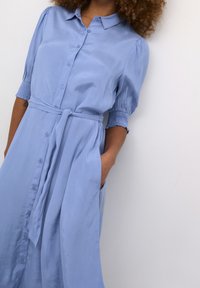 Robe chemise bleu clair en tissu lisse, avec une taille ceinturée, des manches courtes bouffantes et des poches latérales.