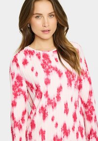 Bluză plisată în roz deschis, cu modele florale roșii, decolteu rotund și mâneci lungi; textură fină și material ușor.