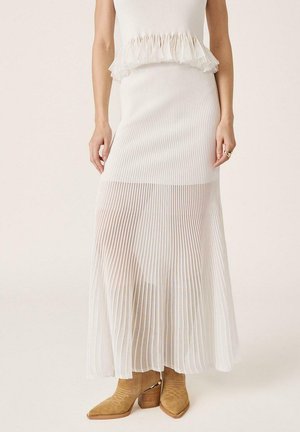 Mujer con una falda larga plisada blanca con detalle de volantes en la cintura, combinada con botines de ante color marrón claro con tacones anchos.