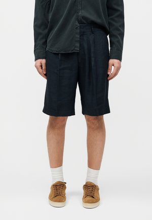 Άνδρας με μαύρο πουκάμισο με κουμπιά, μαύρο σορτς μέχρι το γόνατο, λευκές κάλτσες και καφέ αθλητικά παπούτσια, στέκεται μπροστά σε απλό φόντο.