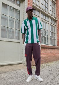Groene en witte gestreepte polo shirt, paarse broek met rode accenten, witte sneakers; staand bij een bakstenen muur en raam.
