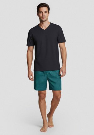 Man staat op blote voeten, draagt een zwart T-shirt met korte mouwen en V-hals en turquoise met patroon bedrukte korte broek tegen een effen achtergrond.