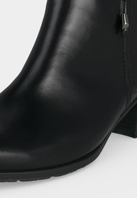 Gros plan d'une botte cheville en cuir noir lisse avec un petit talon bloc et une fermeture éclair latérale.