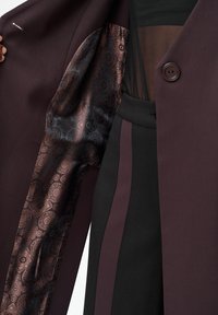 Bordowy płaszcz z wzorzystą ciemną podszewką. Posiada pojedyncze zapięcie na guzik, gładką fakturę, uzupełniony czarnymi i śliwkowymi akcentami.