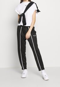 Svarta idrottsbyxor med vita sidskärningar och "NIKE"-logotyp, kombinerat med en vit t-shirt med kort ärm och en svart tröja som hänger över axlarna.