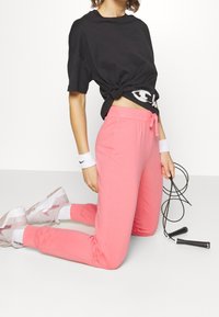 Schwarzes, oversized T-Shirt, am Taillenbund geknotet, kombiniert mit rosa Jogginghosen und weißen Knöchelstrümpfen. Hellrosa Sportschuhe. Springseil in der Hand.