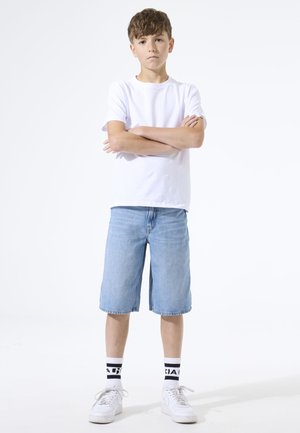 Jongen met lichtbruin haar staat met gekruiste armen, draagt een wit T-shirt, knielange spijkershort, witte sokken met zwarte tekst en witte sneakers.