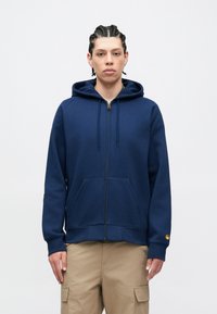 Tamsiai mėlynas užsegamas džemperis su kailiniu gobtuvu, pagamintas iš minkštos medžiagos, su erdvia kengūros kišene, raišteliu gobtuve ir mažu logotipo akcentu ant rankovės.