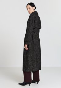 Lungo cappotto nero e grigio con trama, colletto, vita con cintura e tasche laterali, abbinato a pantaloni bordeaux scuro e tacchi neri a punta.