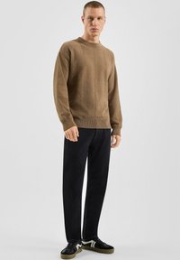 Mężczyzna ubrany w brązowy sweter z fakturą, czarne spodnie i czarne sneakersy z białymi akcentami, stojący na jednolitym jasnym tle.