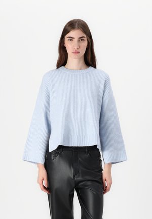 Pull en tricot bleu clair avec un ourlet court et de longues manches larges ; porté sur un pantalons en cuir noir. Texture douce et coupe décontractée.
