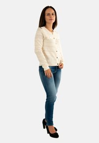 Donna in piedi che indossa un cardigan lavorato a maglia crema con bottoni, jeans blu e décolleté nere, davanti a uno sfondo chiaro e uniforme.