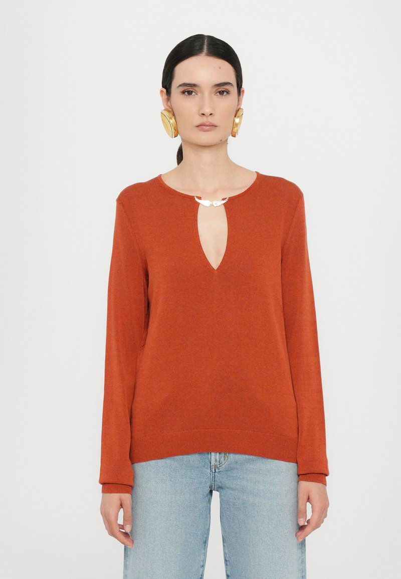 Rust-colored long-sleeve sweater with a keyhole neck detail and silver accent. Soft texture, relaxed fit, paired with light blue jeans.