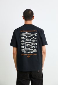 Black cotton t-shirt featuring white fish illustrations and orange text on the back. Design includes label "DICKIES WORKWEAR SINCE '22."