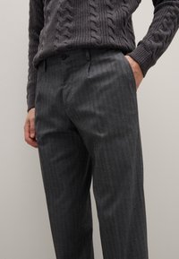 Graue Wollmischhose mit dezentem Nadelstreifenmuster, tailliertem Schnitt, flacher Vorderseite und Seitentaschen. Kombiniert mit einem strukturierten grauen Pullover.