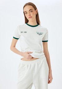 Hvid t-shirt med grøn stribekrave, med teksten "SAN DIEGO" og "EST. 1992" trykt på. Båret med lyse sweatpants.