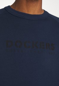Námořnická modrá mikina vyrobená z měkké tkaniny, s kulatým výstřihem a výrazným černým potiskem, který zní "DOCKERS ESTABLISHED 1986."
