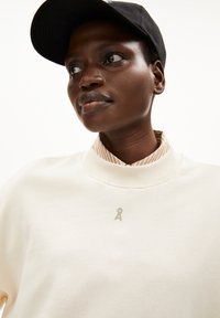 Crème sweatshirt met hoge kraag, voorzien van een subtiel logo op de borst. In combinatie met een gestreepte kraagshirt eronder en een zwarte pet.