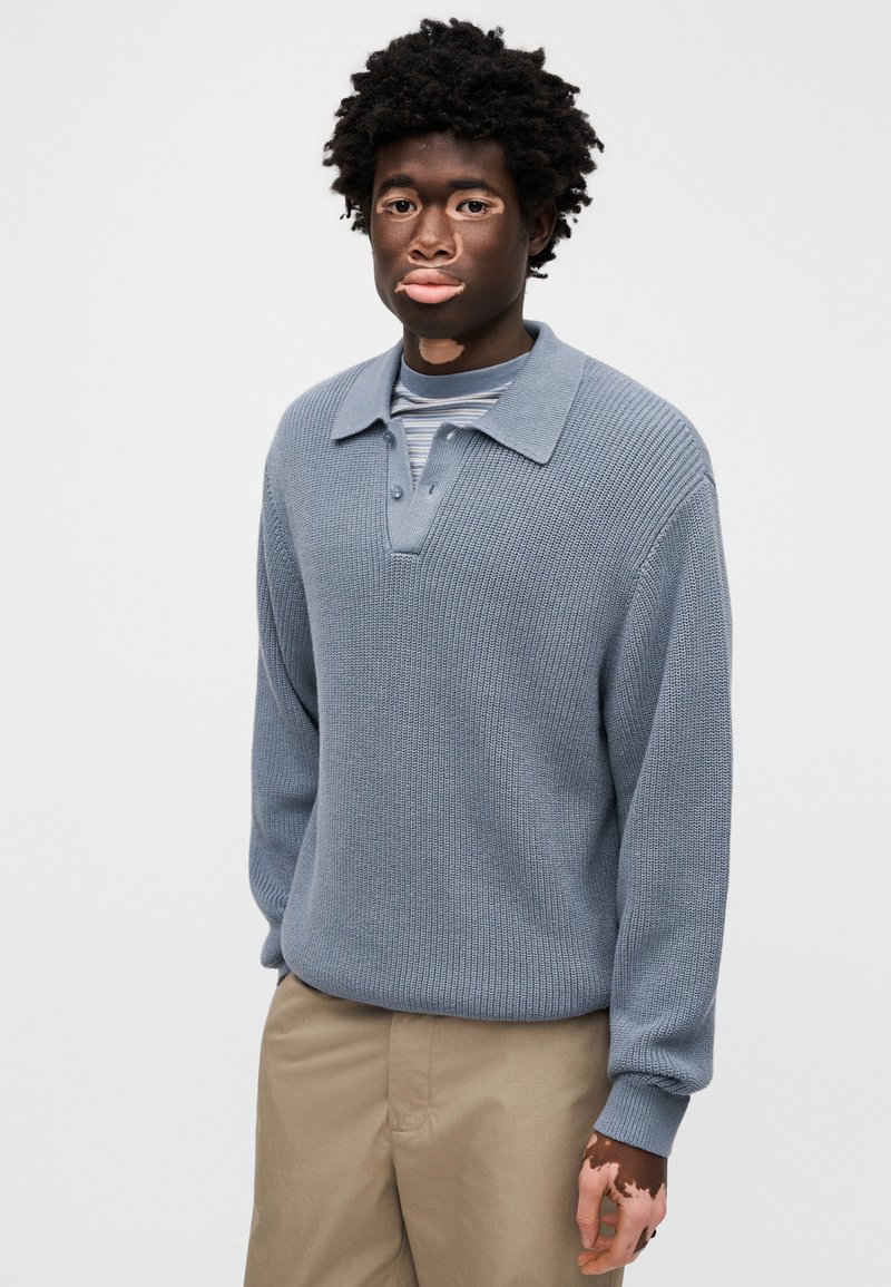 Strikket, lyseblå pullover med krave og knapstolpe. Lange ærmer med ribkant og tekstureret stof. Båret over en stribet skjorte.