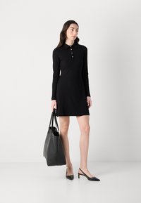 Zwarte lange mouwen jurk met een kraag en knoopdetails, gecombineerd met een zwarte tote en puntige slingback schoenen, voor een effen achtergrond.