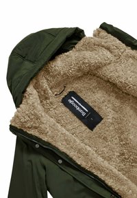 Giacca esterna verde con un rivestimento interno in pile beige, morbido e texturizzato. Presenta un cappuccio, una zip e un'etichetta di branding all'interno.