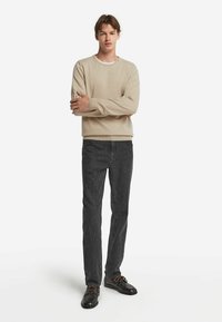 Beige stickad tröja med rund halsringning, kombinerad med mörkgrå avsmalnande jeans och svarta skor, med bruna snören och subtil textur.