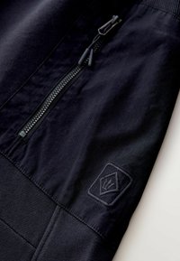 Mustast sportjope sektsioon, kus on lukuga tasku ja tekstureeritud kangas. Küljel on teemantses kujus reljeefne logo.