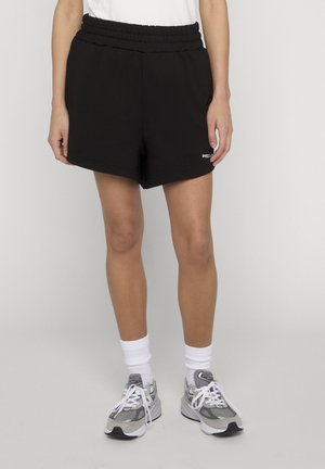 Person iført sorte elastiske talje-shorts, hvide sokker og grå og hvide sneakers, står mod en ensfarvet baggrund.