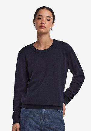 Kvinde iført en mørkeblå crewneck-trøje og blå jeans, poserer med den ene hånd på hoften mod en hvid ensfarvet baggrund.