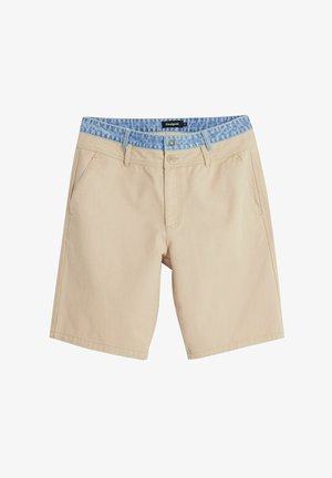 Pantalones cortos beige con una cinturilla de patrón azul similar al denim. Presenta bolsillos frontales, una cremallera y detalles de costura en contraste.