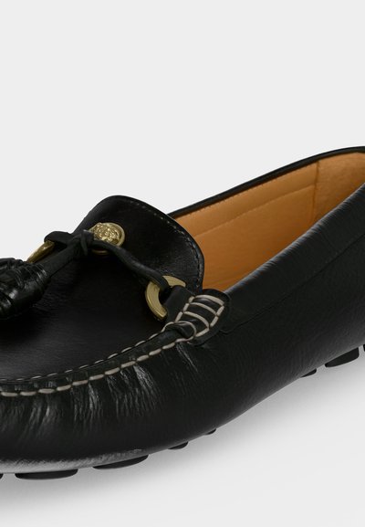 Mocassin en cuir noir avec coutures blanches, œillets métalliques dorés et détail de lacet en cuir noué, avec une doublure intérieure beige.