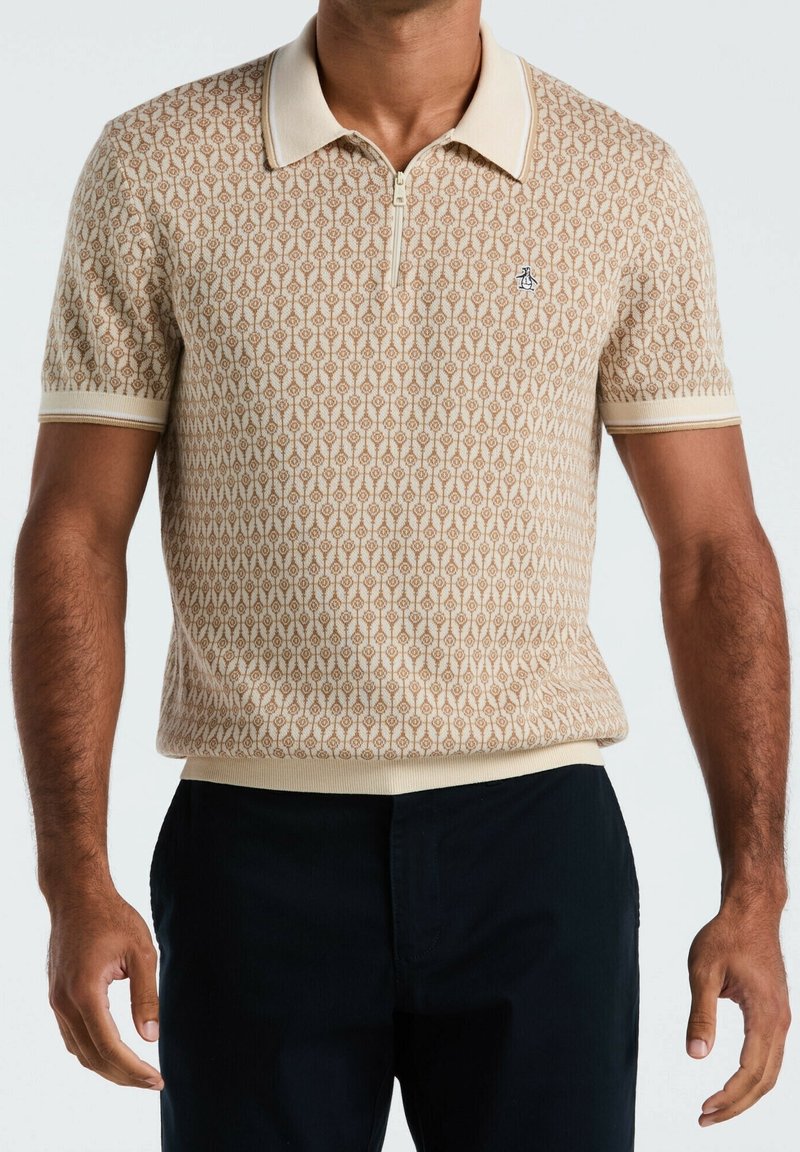 Homme portant un polo beige à manches courtes à motif, avec un col zippé et un petit logo sur la poitrine, associé à un pantalon noir.
