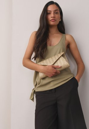 Žena s dugom tamnom kosom u maslinasto zelenu majicu bez rukava i crne hlače, držeći presavijenu tkanu torbicu ispred običnog zida.