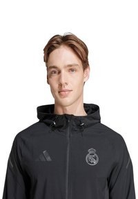 Mustapuku, jossa on huppu, vetoketjukiinnitys, teksturoitu kangas, Adidas-logo ja Real Madridin emblem vasemmassa rintapielessä.