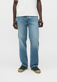 Ανοιχτό μπλε τζιν με ίσια γραμμή στα πόδια, κατασκευασμένα από denim. Φορεμένα με πράσινα αθλητικά παπούτσια που διαθέτουν λαστιχένια σόλα. Απλός σχεδιασμός χωρίς ορατές φθορές.