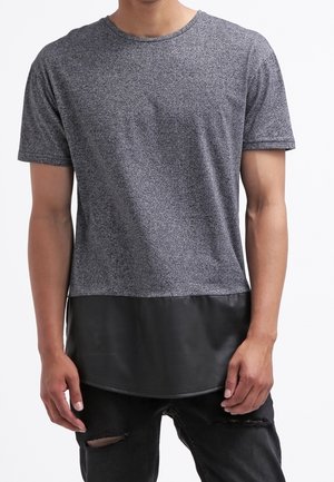 Mężczyzna ubrany w szarą koszulkę z krótkim rękawem z błyszczącym, czarnym, warstwowym wykończeniem oraz czarne podniszczone dżinsy, stojący na tle białego tła.