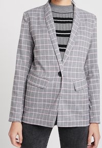 Šedý kostkovaný blazer se střihovým přizpůsobením, který má klopový límec, dvě přední kapsy a jednobodové zapínání. Texturovaná tkanina a detaily vzoru.