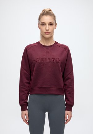 Жена представя бордо суичър GUESS със сив клин на изчистен бял фон.